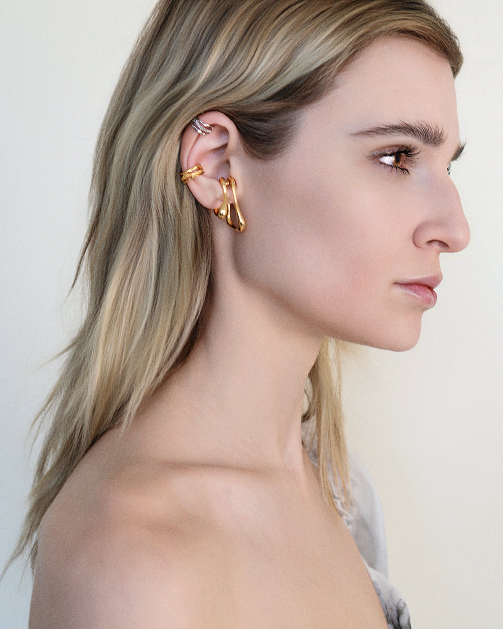 Model wearing oversized clip-on earrings in gold and silver versions. These sculptural, teardrop-shaped earrings feature a smooth, fluid form that wraps elegantly around the earlobe. Designed for non-pierced ears, they offer a bold, modern statement with a comfortable fit. Perfect for elevating everyday looks or adding a refined touch to evening outfits.