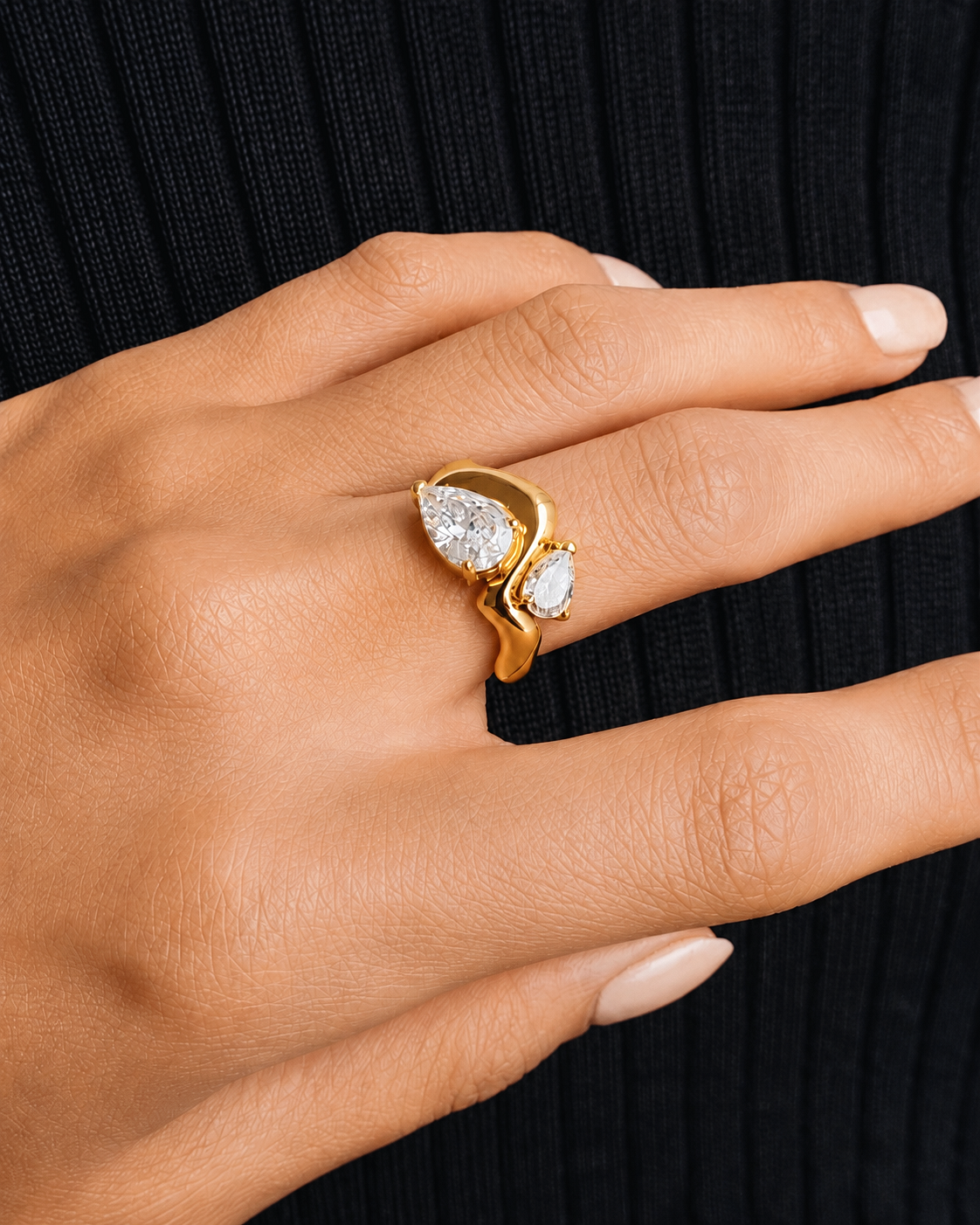 A close-up of a hand wearing a gold ring with two pear-cut clear gemstones, set against a black ribbed knit background.