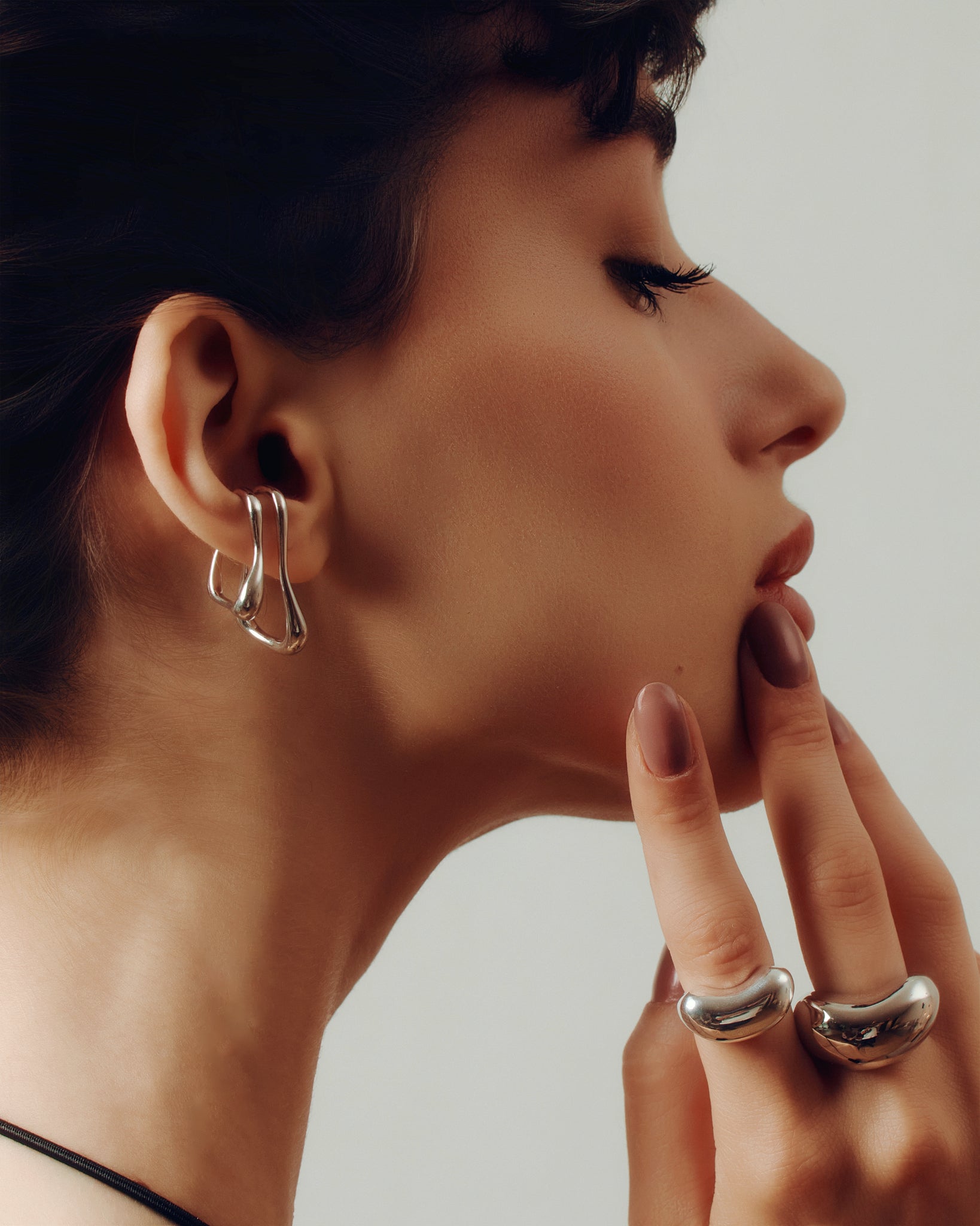 Model wearing oversized clip-on earrings in gold and silver versions. These sculptural, teardrop-shaped earrings feature a smooth, fluid form that wraps elegantly around the earlobe. Designed for non-pierced ears, they offer a bold, modern statement with a comfortable fit. Perfect for elevating everyday looks or adding a refined touch to evening outfits.