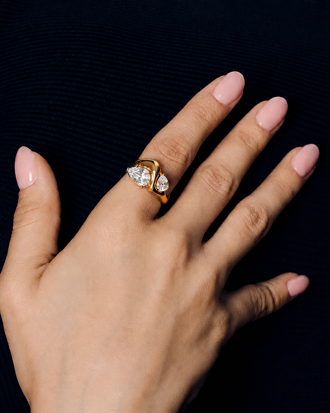 A close-up of a hand wearing a gold ring with two pear-cut clear gemstones, set against a dark textured background.