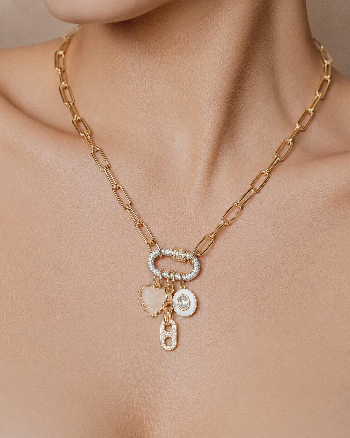 A close-up of a gold chain necklace worn on a model's neck, featuring a central oval link with a pavé-set clear stone and dangling charms including a pale pink heart, a round clear stone charm, and a small gold initial 'H' charm.