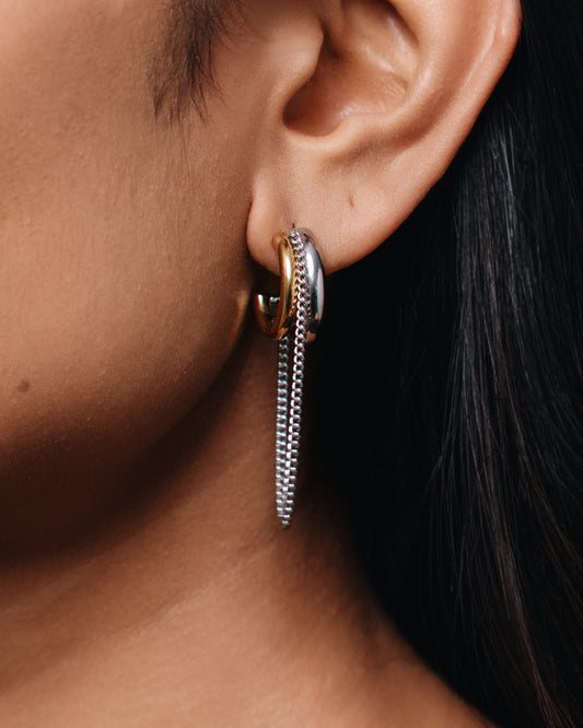 Two-tone gold and silver hoop earrings worn on the ear, featuring a mixed-metal hoop with a long chain dangle detail.