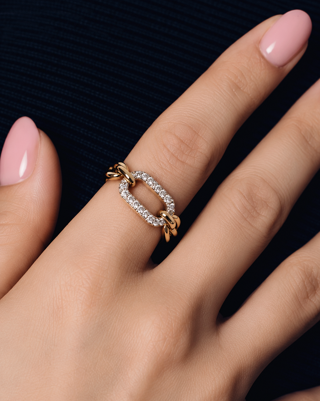 A close-up of a hand wearing a gold ring with a diamond-set oval center and twisted gold side details, against a dark navy ribbed background.
