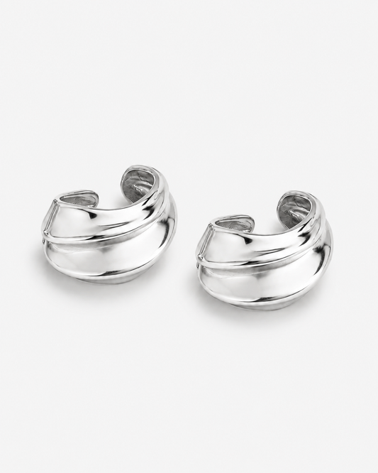 A pair of polished silver huggie hoop earrings with a modern, layered curved design, displayed on a clean light gray background.