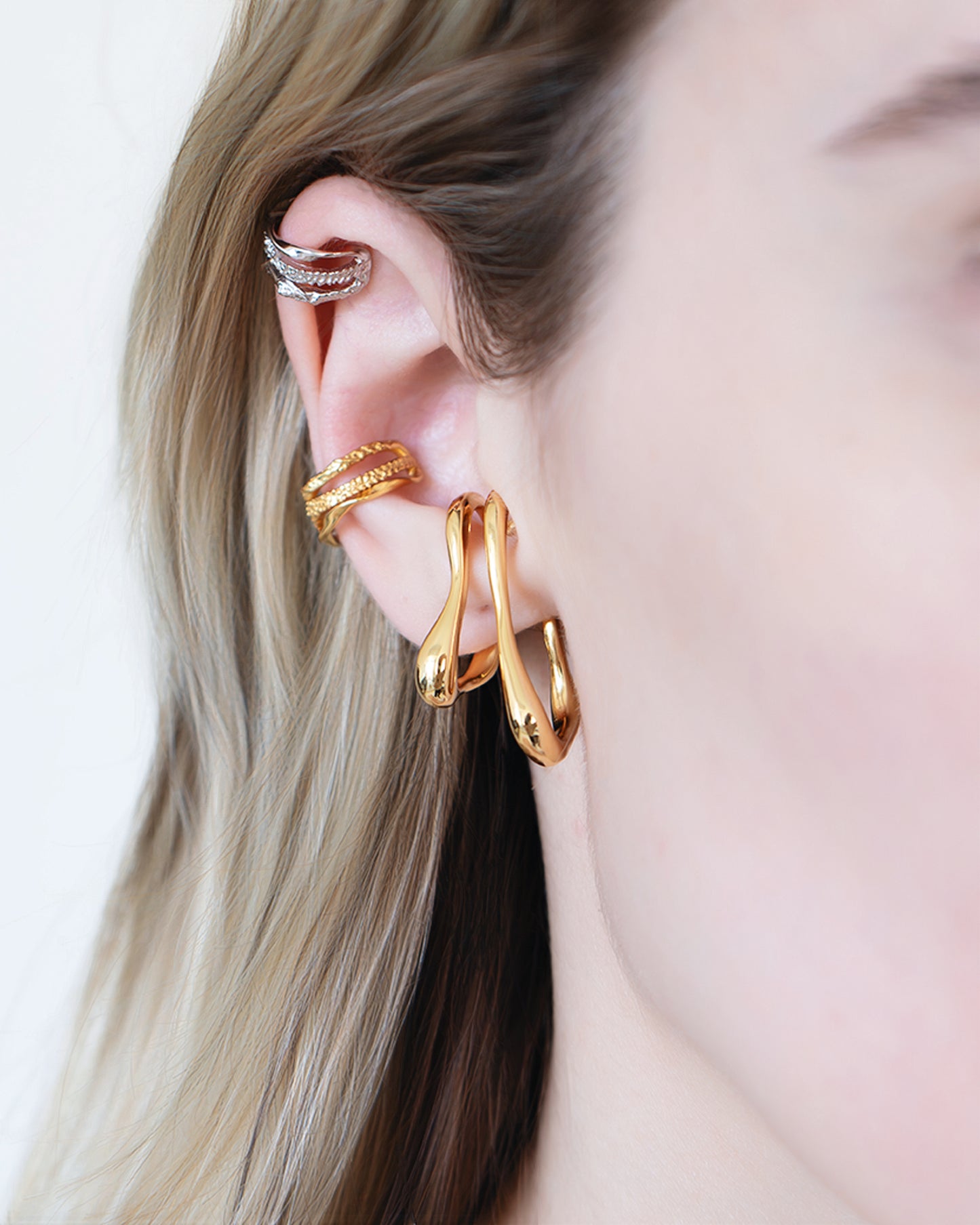 Model wearing oversized clip-on earrings in gold and silver versions. These sculptural, teardrop-shaped earrings feature a smooth, fluid form that wraps elegantly around the earlobe. Designed for non-pierced ears, they offer a bold, modern statement with a comfortable fit. Perfect for elevating everyday looks or adding a refined touch to evening outfits.