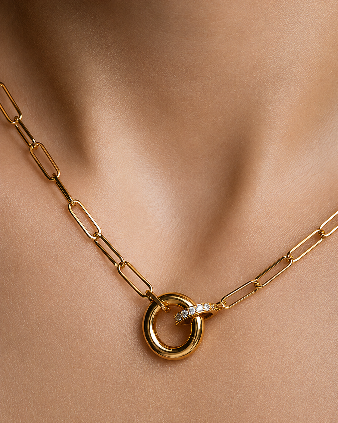 Gold paperclip chain necklace with interlocking circular pendant and a small pavé diamond accent, shown worn on a model's neck.
