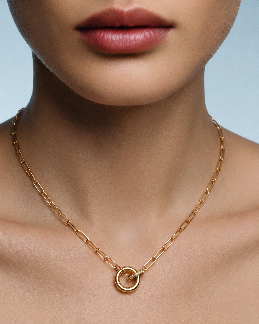 A close-up of a model's neck and lower face wearing a delicate gold paperclip chain necklace with a small interlocking circle zircon charms pendant.