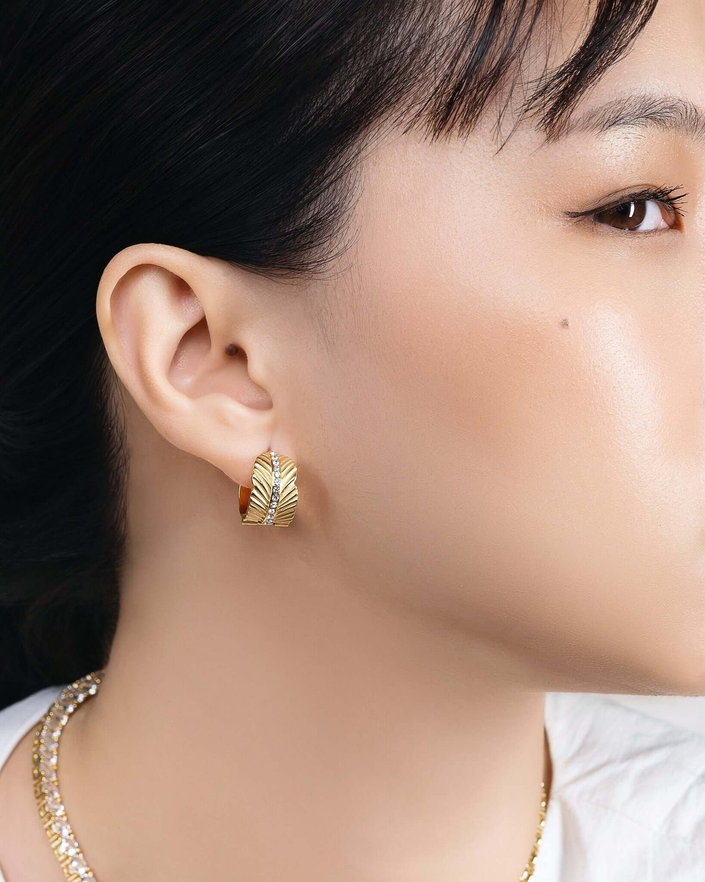 Woman wearing chunky gold CZ hoop earrings with textured sunburst pattern and crystal center stripe, statement gold huggie hoop earrings for women, trendy luxury gold earrings, modern fashion jewelry, bold everyday hoop earrings, stylish gold crystal earrings model closeup.