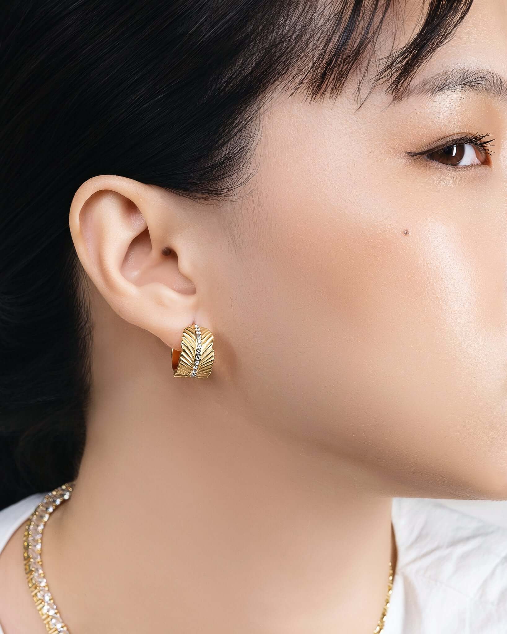 Woman wearing chunky gold CZ hoop earrings with textured sunburst pattern and crystal center stripe, statement gold huggie hoop earrings for women, trendy luxury gold earrings, modern fashion jewelry, bold everyday hoop earrings, stylish gold crystal earrings model closeup.