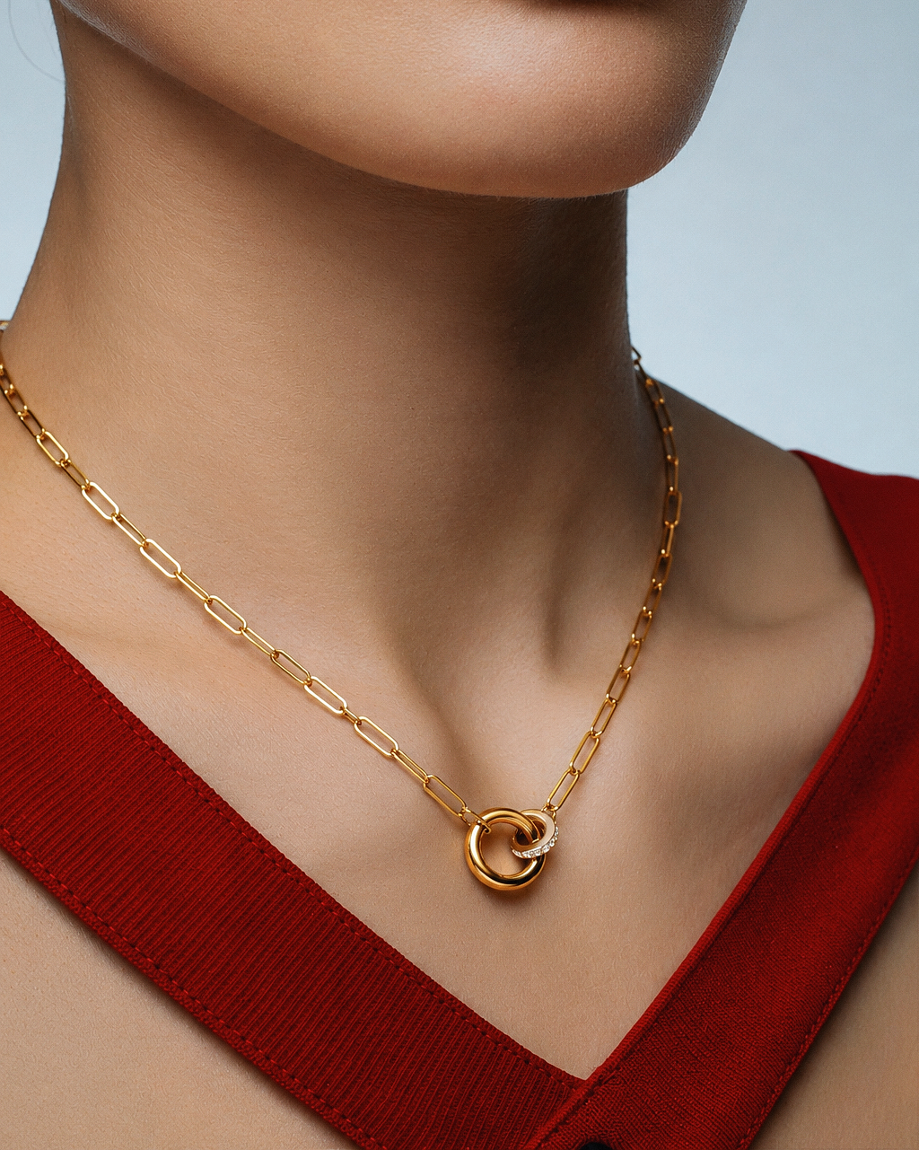 A close-up of a model's neck and chest wearing a gold paperclip chain necklace with a double-ring pendant, paired with a red ribbed top against a light gray background.