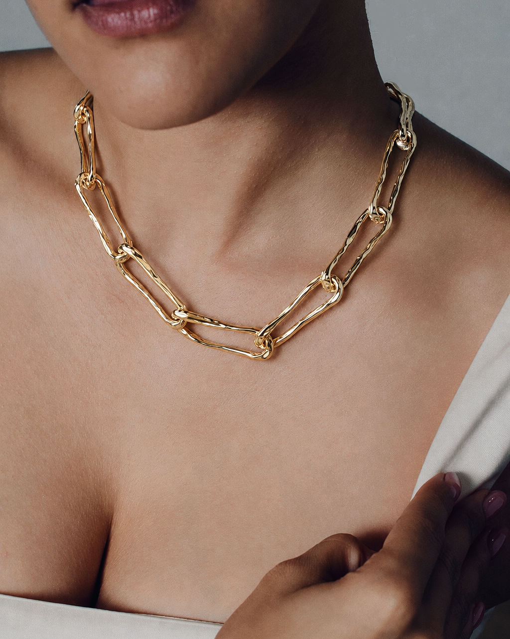 A close-up of a model wearing a chunky gold chain necklace with interlocking rectangular links, styled with a white strapless top against a neutral gray background.