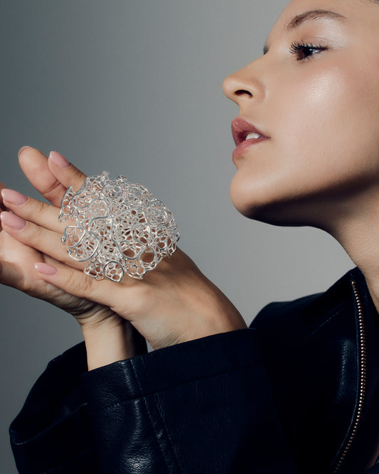 Close-up of a sculptural open-work statement ring featuring organic, lattice-like curves that flow across the fingers. The bold, artistic design catches light and shadow, creating a striking balance of size and airiness. Perfect for modern, contemporary, and architectural jewelry lovers who appreciate wearable art.