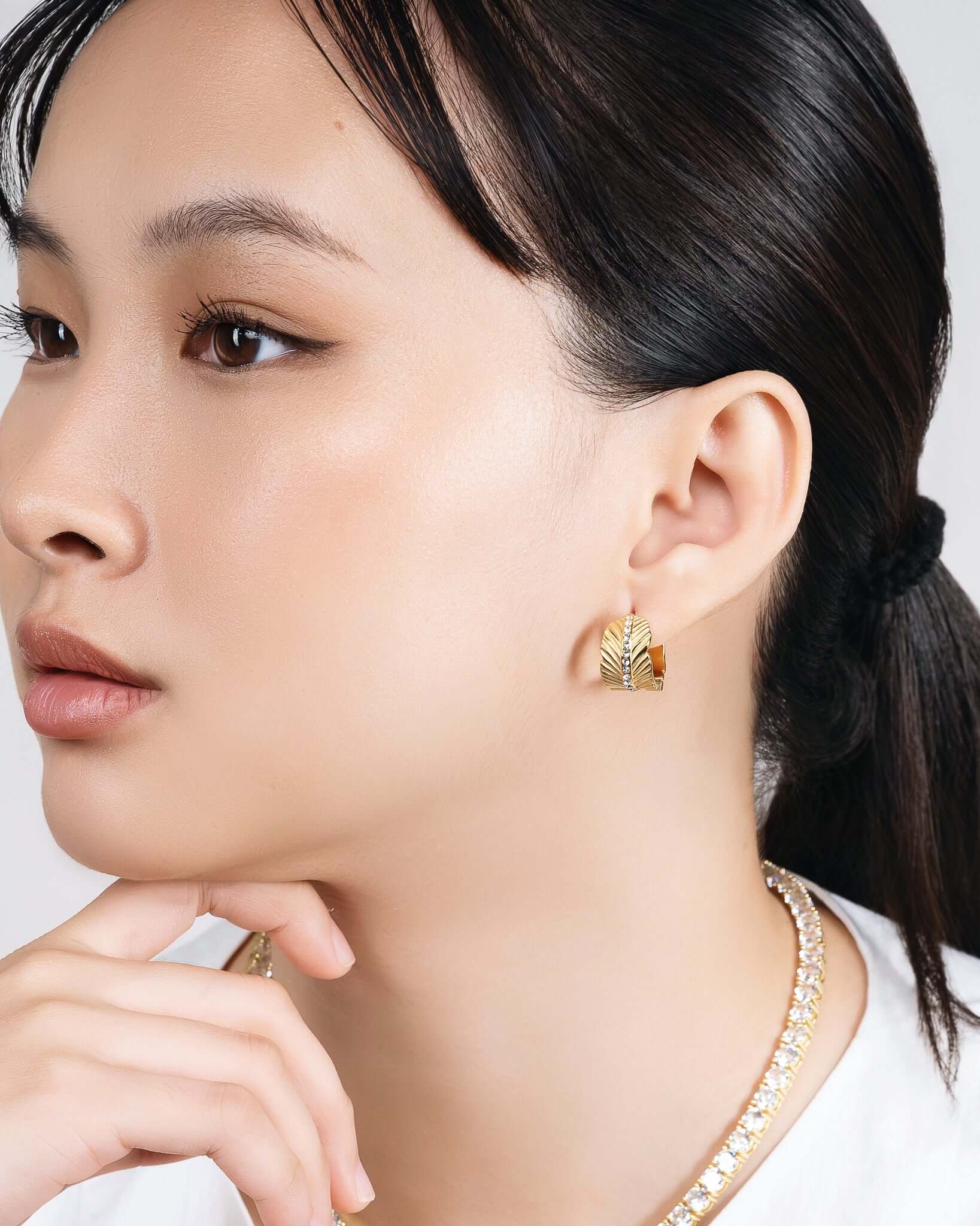 Woman wearing chunky gold CZ hoop earrings with textured sunburst pattern and crystal center stripe, statement gold huggie hoop earrings for women, trendy luxury gold earrings, modern fashion jewelry, bold everyday hoop earrings, stylish gold crystal earrings model closeup.