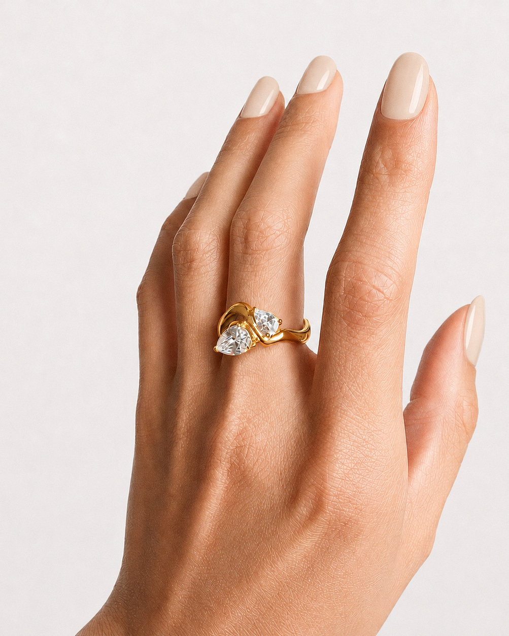 A close-up of a hand wearing a gold ring with two pear-cut clear gemstones, set against a clean light gray background.