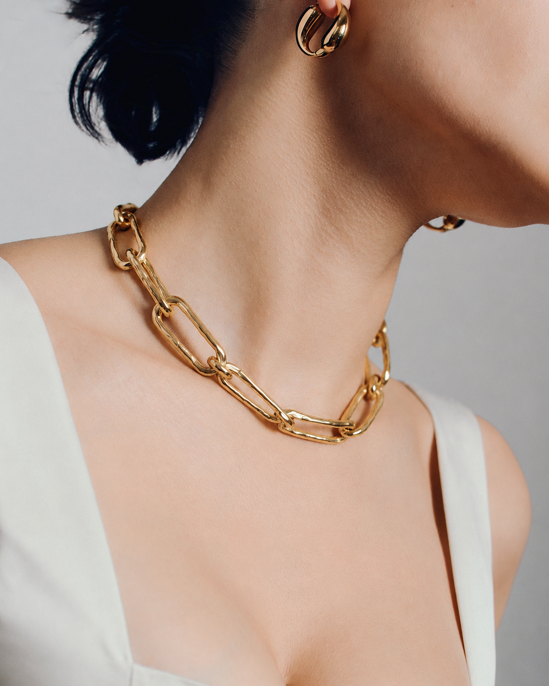 A close-up of a model wearing a chunky gold chain necklace with oversized links, styled with matching gold hoop earrings and a white top against a neutral gray background.