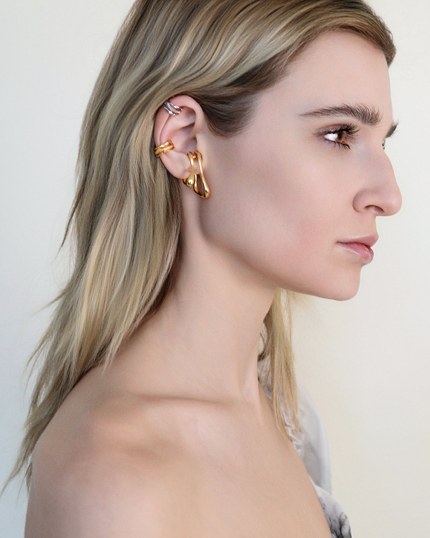 Model wearing oversized clip-on earrings in gold and silver versions. These sculptural, teardrop-shaped earrings feature a smooth, fluid form that wraps elegantly around the earlobe. Designed for non-pierced ears, they offer a bold, modern statement with a comfortable fit. Perfect for elevating everyday looks or adding a refined touch to evening outfits.