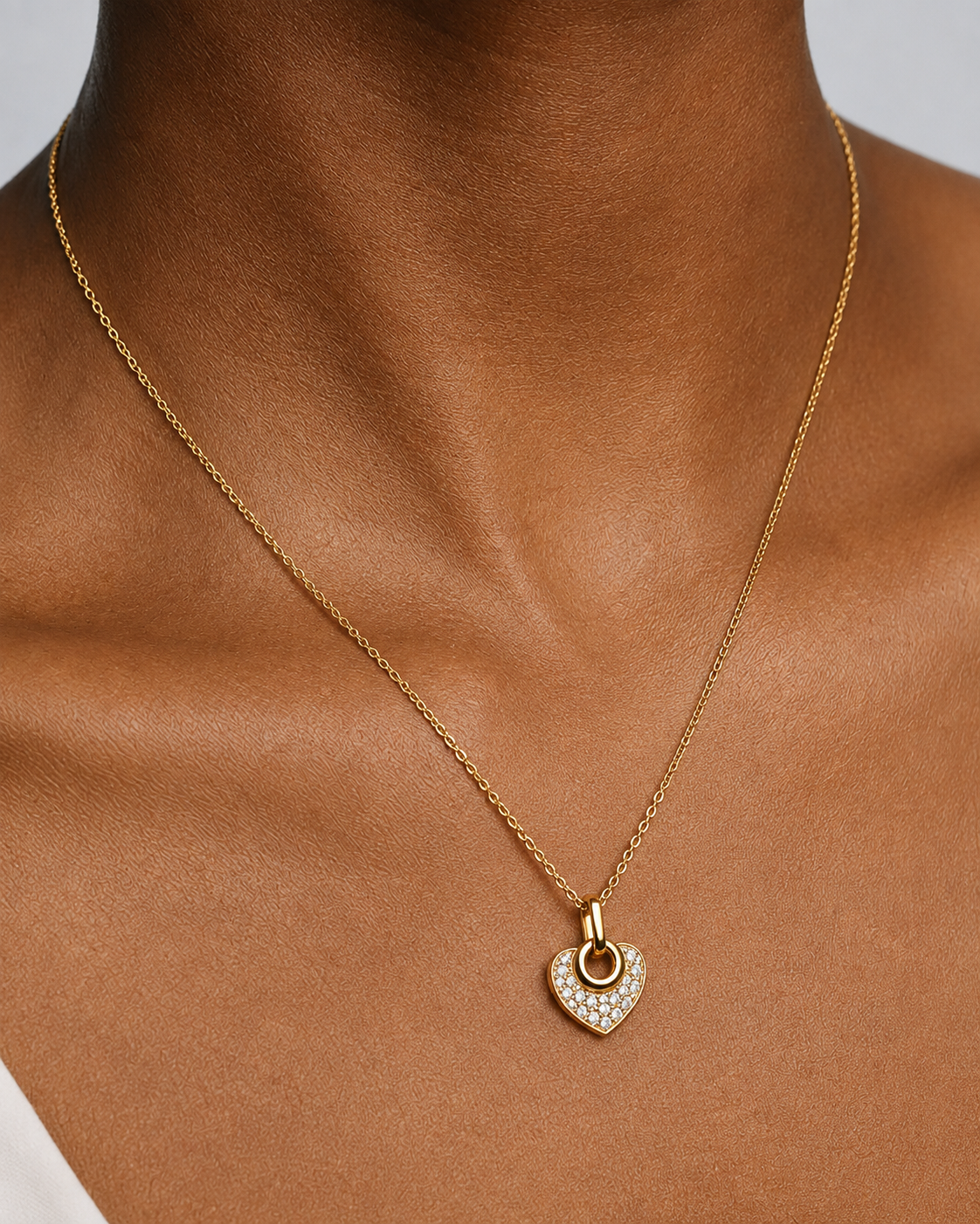 A gold chain necklace with a small heart-shaped pendant encrusted with clear stones, shown worn on a model's neck.