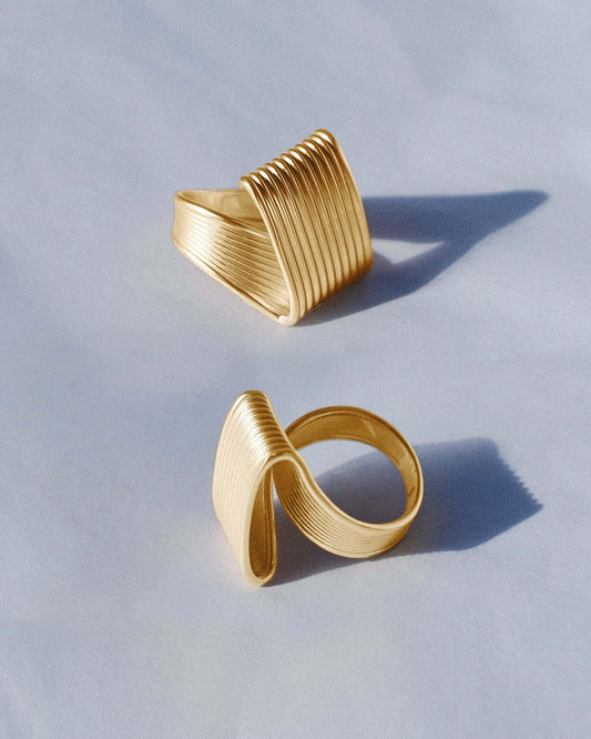 Pleated square gold ring featuring fine ridged detailing that flows seamlessly across the face and band. This sculptural textured ring combines a bold square shape with a soft pleated design, balancing modern geometry and fluid form. Crafted in polished gold-tone metal, it’s a statement piece for everyday or occasion wear.