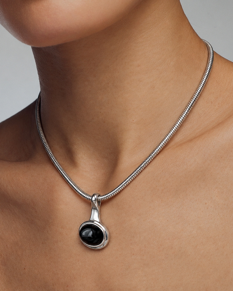 A close-up of a model's neck and chest wearing a silver snake chain necklace with a round black onyx pendant.