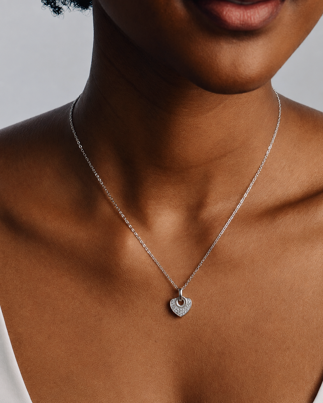 A close-up of a model wearing a delicate silver chain necklace with a small, openwork heart-shaped pendant set with tiny clear stones, shown against the model's neck and upper chest.