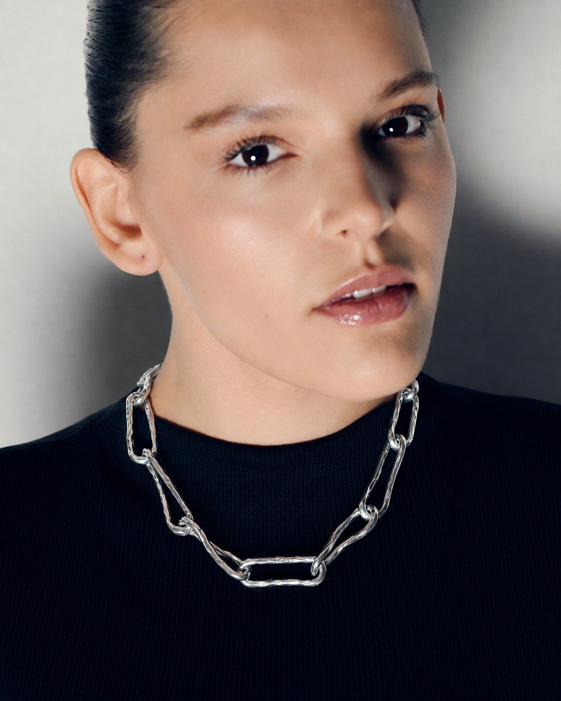 A close-up portrait of a model wearing a chunky silver chain-link necklace with elongated links, paired with a black ribbed top against a neutral gray background.