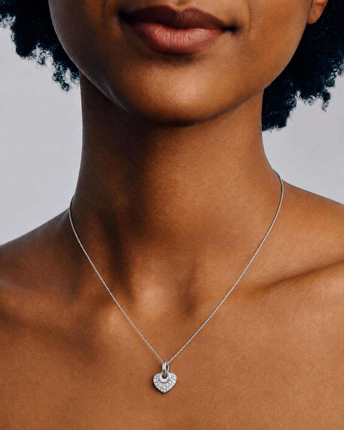 A close-up of a model's neck and chest wearing a delicate silver chain necklace with a small, heart-shaped pavé pendant.