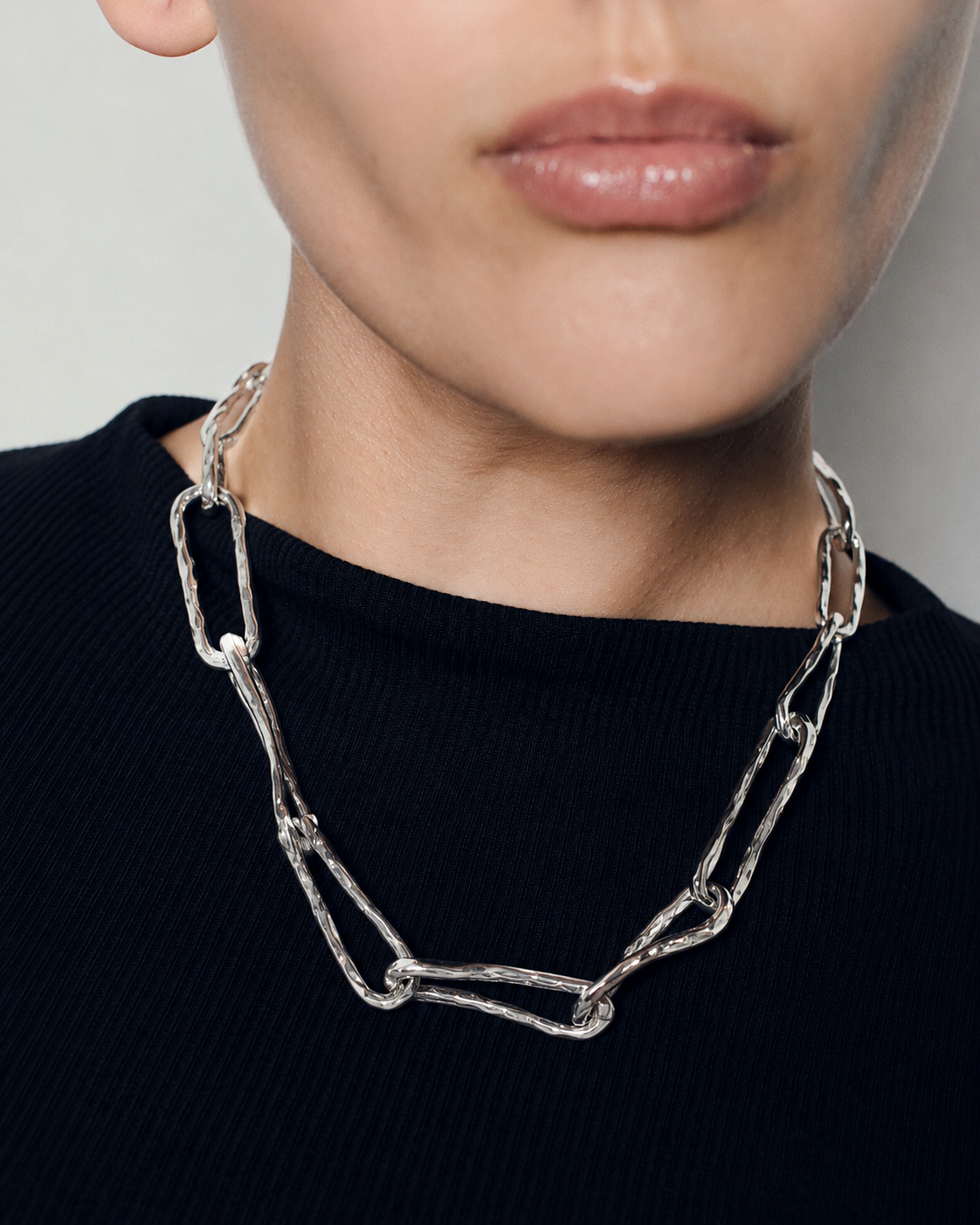 A close-up of a model wearing a chunky, textured silver chain-link necklace over a black ribbed top.
