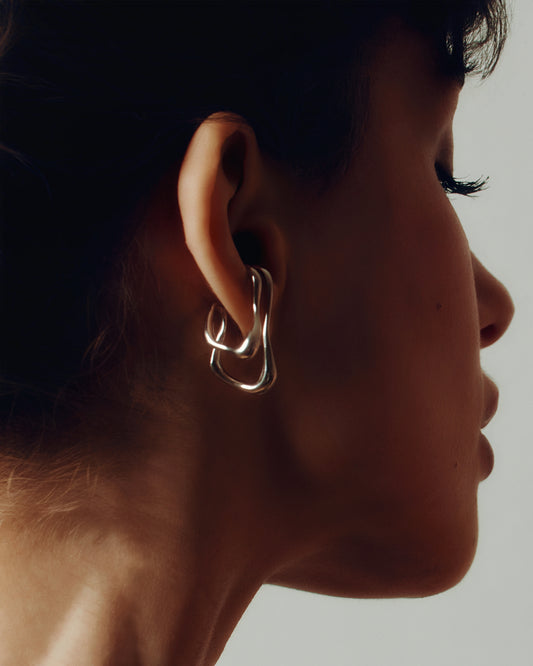 Model wearing oversized clip-on earrings in gold and silver versions. These sculptural, teardrop-shaped earrings feature a smooth, fluid form that wraps elegantly around the earlobe. Designed for non-pierced ears, they offer a bold, modern statement with a comfortable fit. Perfect for elevating everyday looks or adding a refined touch to evening outfits.