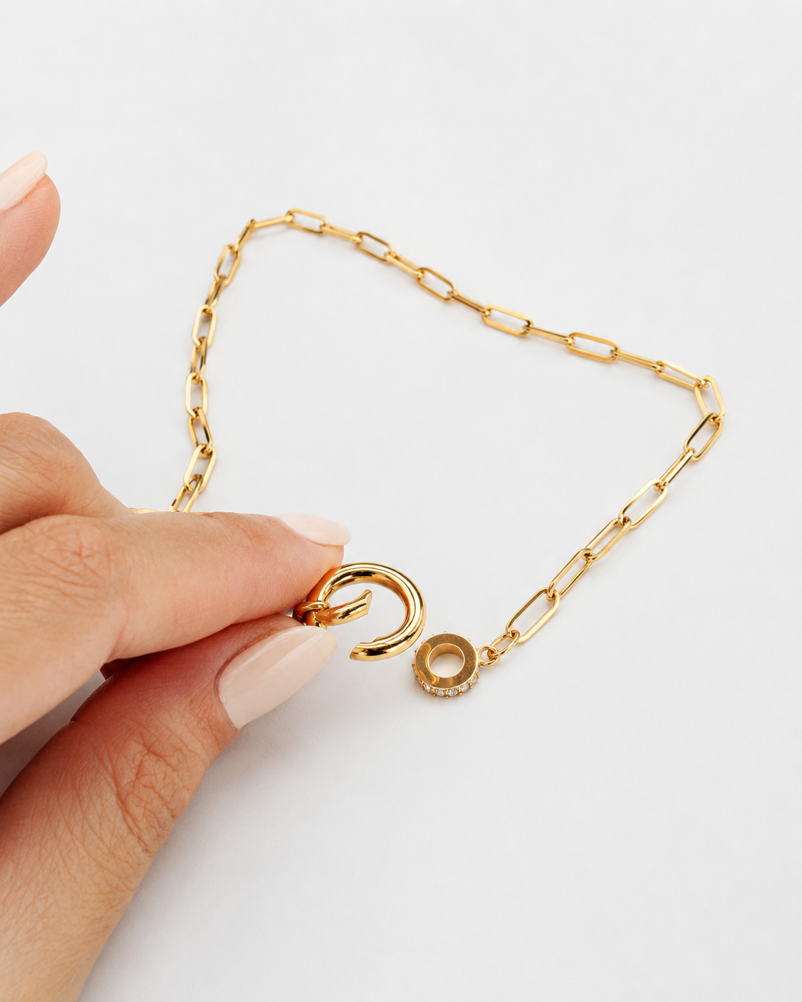 A gold paperclip chain necklace with a small circular charm, held by a hand against a clean white background.