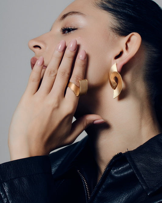 Pleated sculptural gold earrings featuring a twisted, folded design with fine ridged texture. These bold statement earrings have an open curved form that adds dimension and movement. Crafted in polished gold-tone metal, they bring a modern, architectural edge to any outfit — lightweight yet striking for everyday or occasion wear.