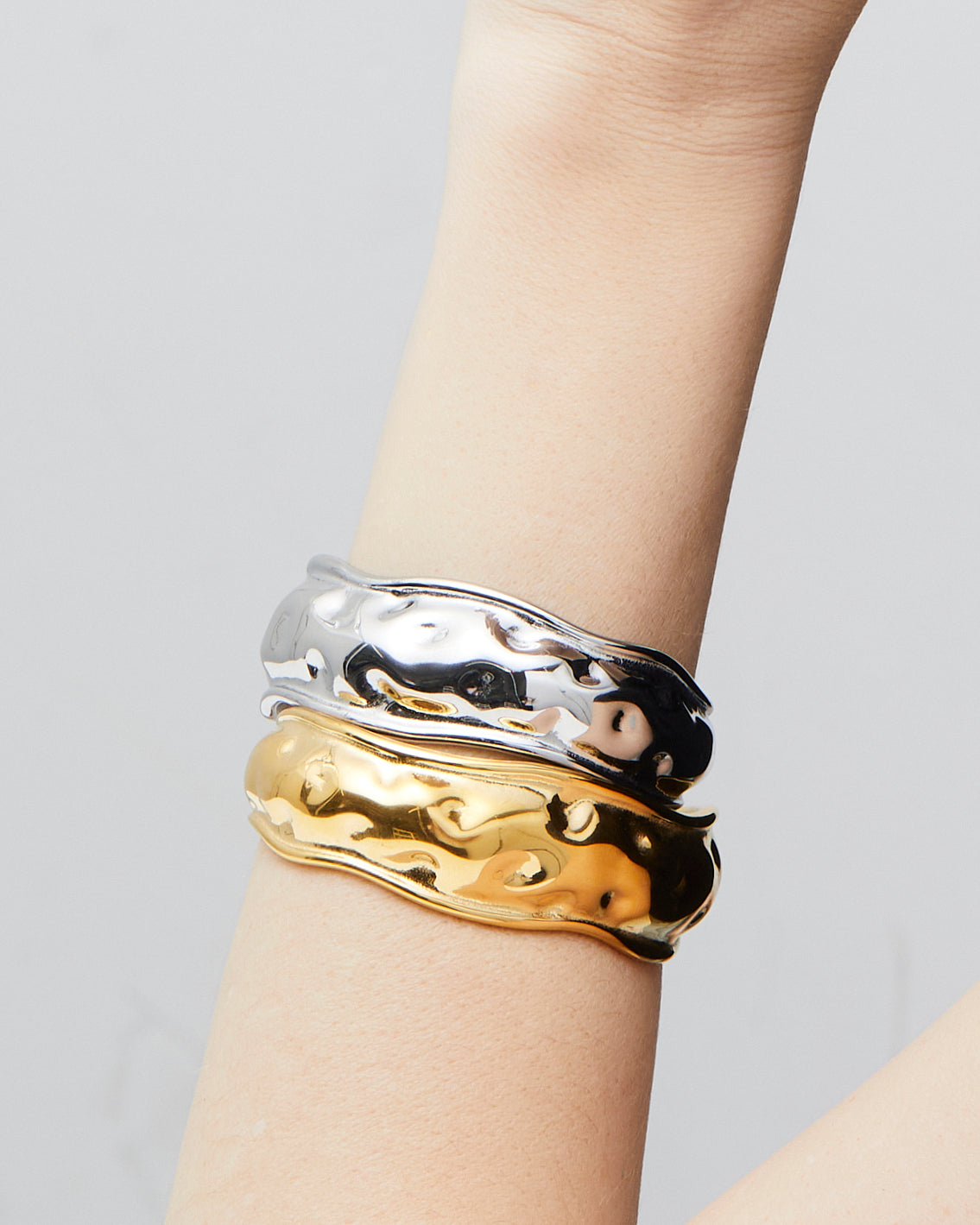 Gold or silver wavy cuff bracelets stacked on wrist, featuring a sculptural organic design with high-polish mirror finish and hammered texture — modern statement bangles crafted from durable Italian metal.